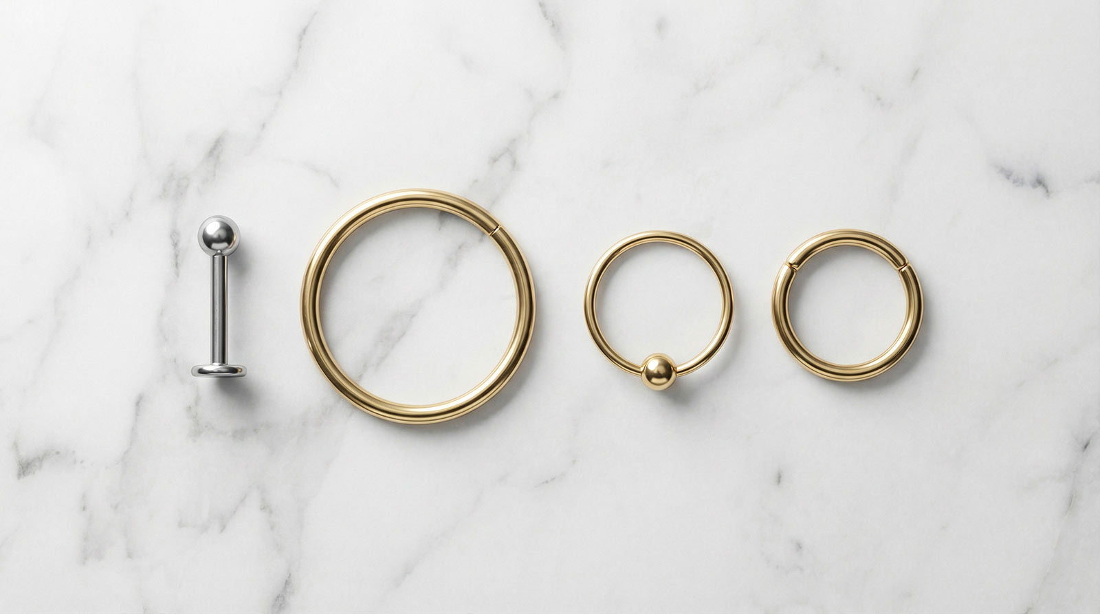 Conch piercing jewelry options — titanium flat-back labret stud, large gold cartilage hoop 12mm, orbital conch hoop and clicker ring on white marble