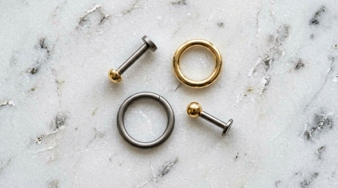 Tragus piercing jewelry flatlay — titanium flat-back labret stud, gold huggie hoop 6mm, seamless titanium hoop and threadless push-pin tragus earrings on white marble